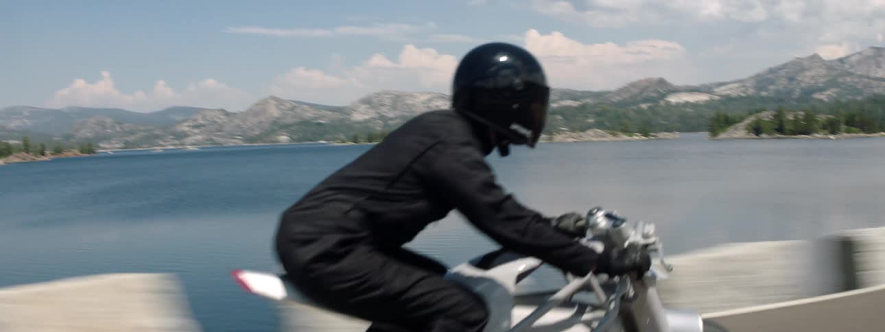 Motorcyclist rides along highway across lake from snowy mountains