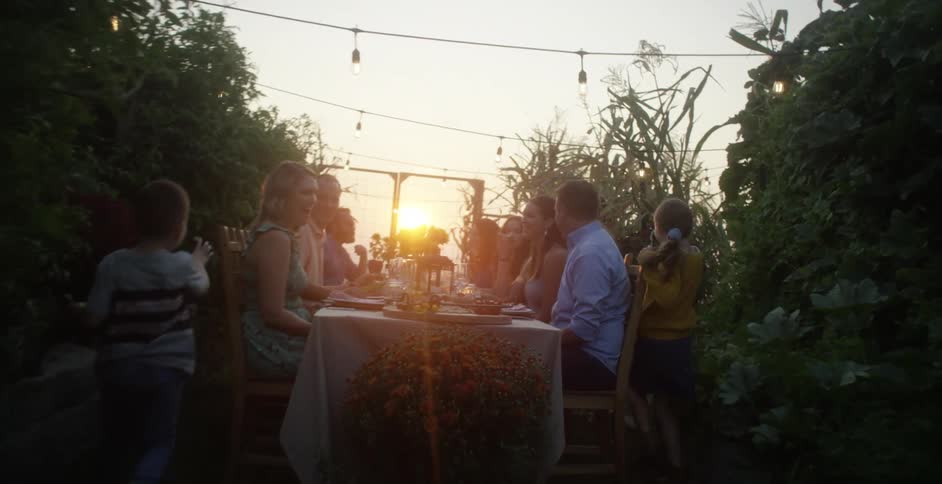 Family and friends eat at long table in garden as sun sits low in red sky