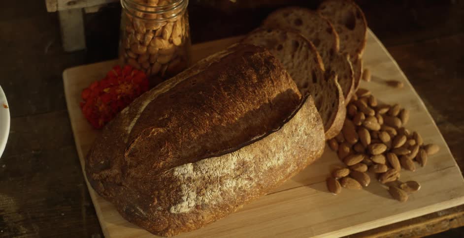 Bread that is partially sliced on a cutting board with nuts next to it on the board and in a jar