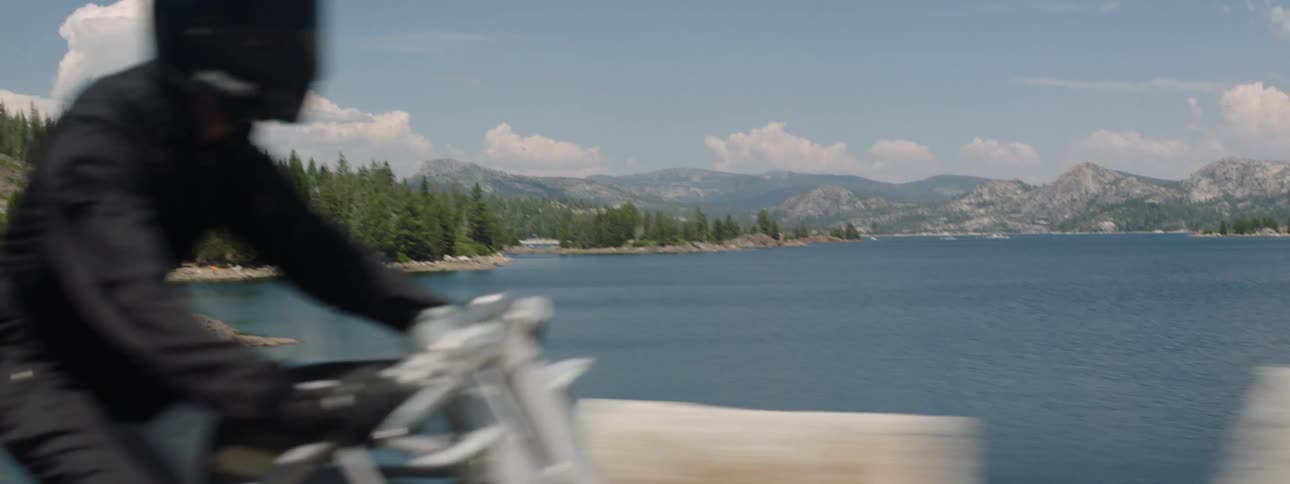 A motorcycle travels on a bridge over a lake with forests and mountains in the distance as the rider is wearing a black jacket and helmet