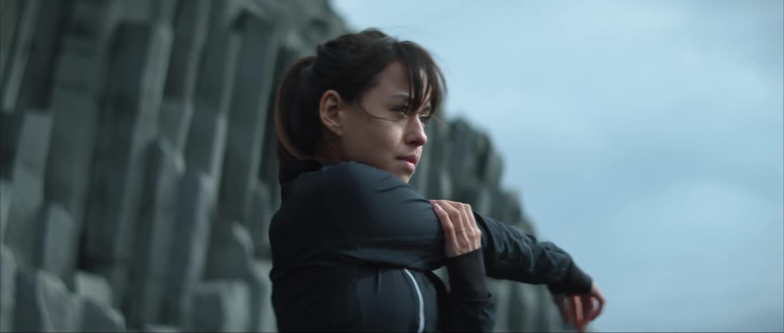 A woman in a black jacket stands outdoors and stretches her arm across her chest