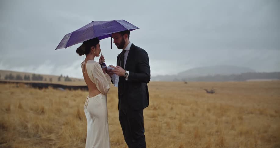 Groom reads from paper while he and bride stand under umbrella in field while it rains