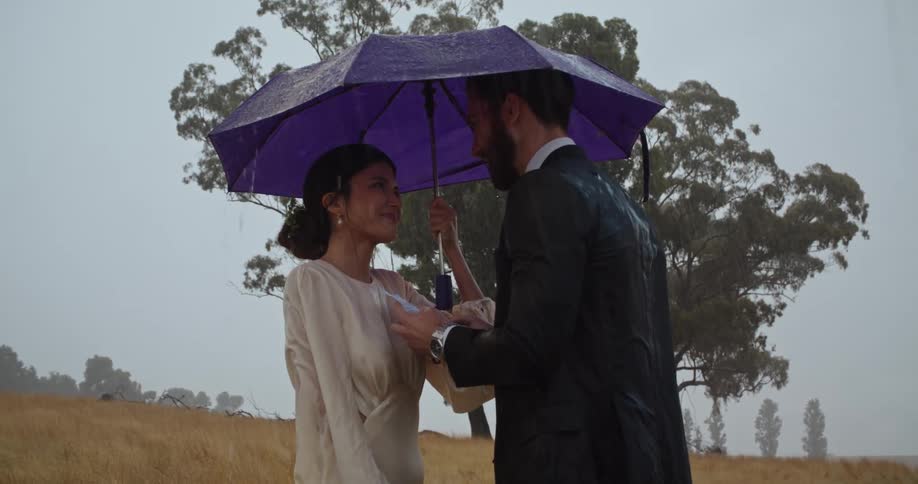 A man and woman stand outside under an umbrella as the man is talking