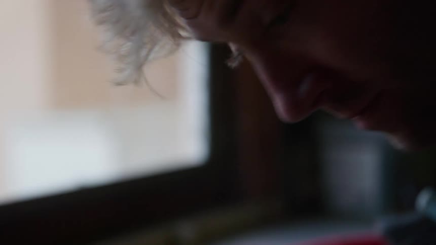 A man blows on something while standing by his kitchen window