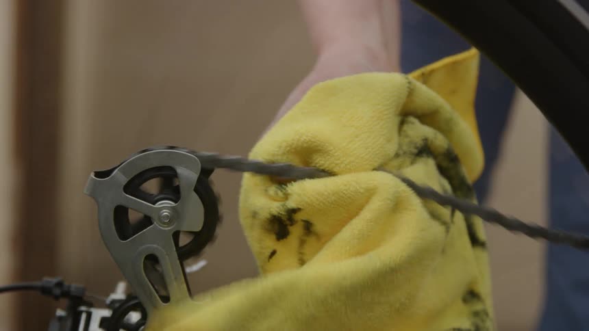 A man is turning a bicycle peddle and holding a yellow cloth on the chain to clean it as it moves through the rear derailleur