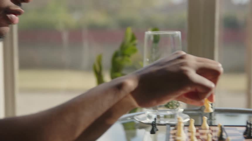 A man is sitting at a glass table playing chess near a window as he moves a game piece his opponent reaches their hand across the game board