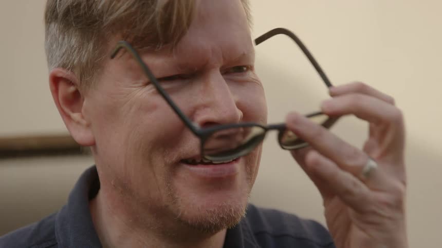 A man wipes his forehead and smiles as he puts on his sunglasses
