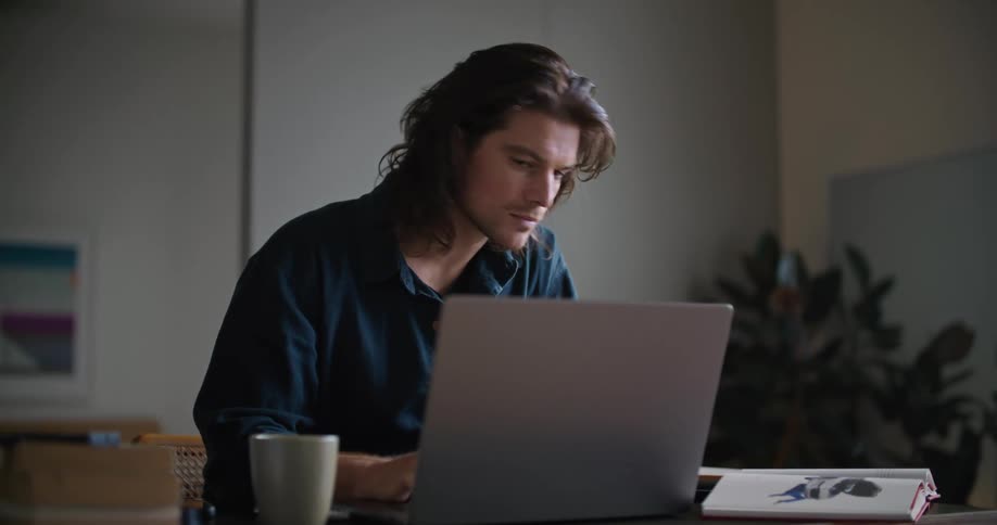 Man sitting at desk with laptop alternating between typing on keyboard and referencing book on desk