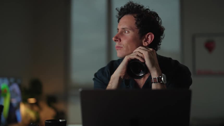A man sits at his laptop while holding a camera lens and pondering a decision
