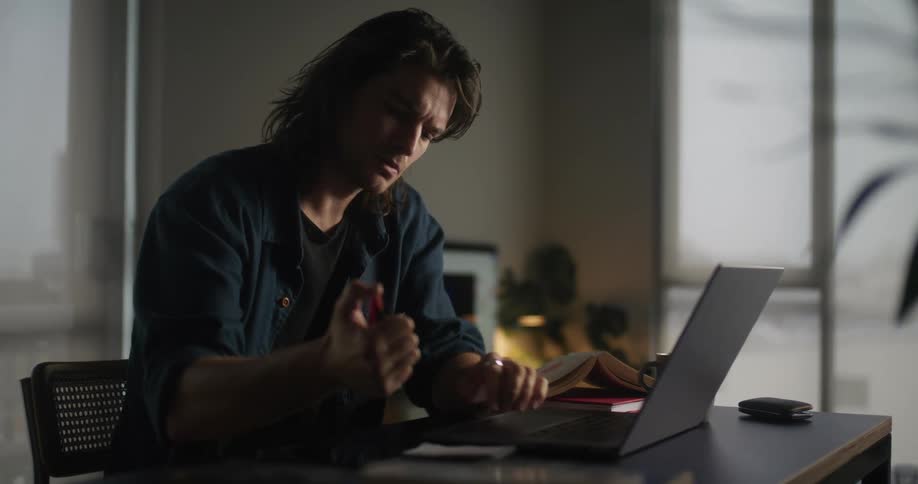 A man sitting at his desk typing on a laptop and then writing on a notepad
