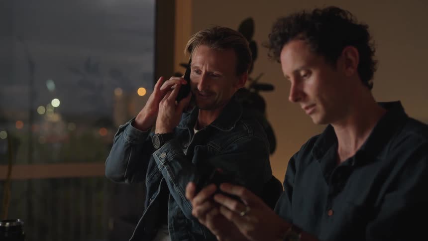 Two men sitting next to each other one talking on the phone while the other holds a camera lens