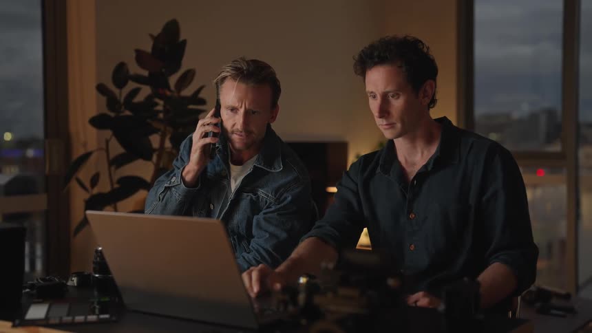 Two men sitting next to each other in front of a laptop one on the phone and both looking at the laptop