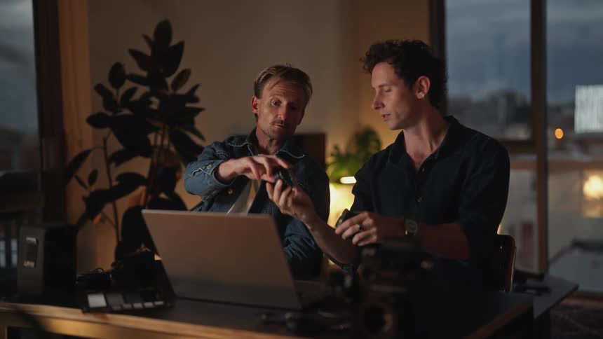 Two men sitting next to each other holding and looking at camera lenses in front of a laptop