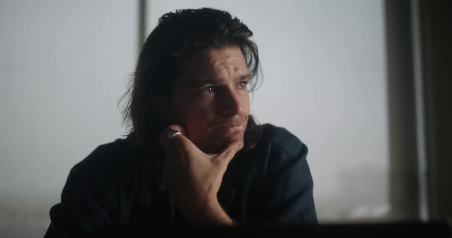 A man sitting and reading something in front of him then putting his hand on his chin and looking contemplatively