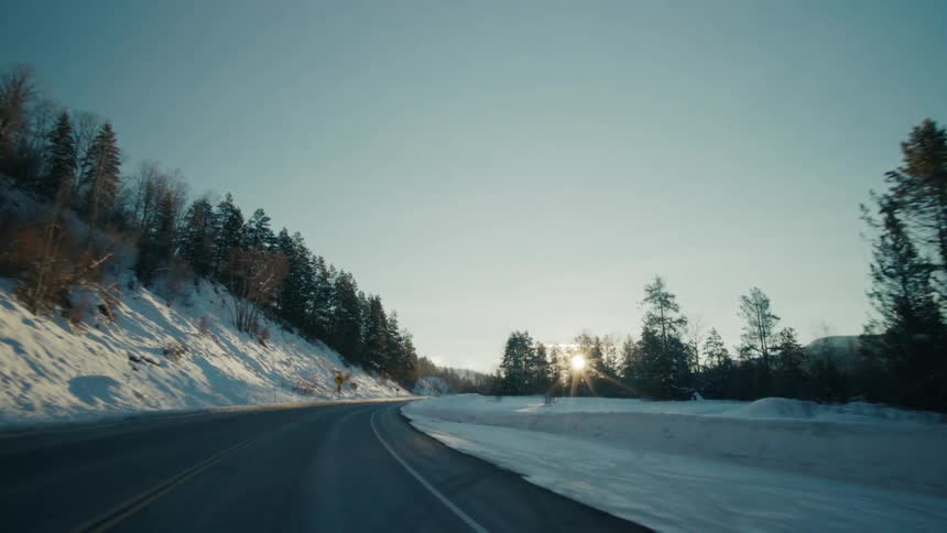 Traveling around a curve on a rural highway with snow and trees on the roadside and the sun shining brightly over mountains in the distance