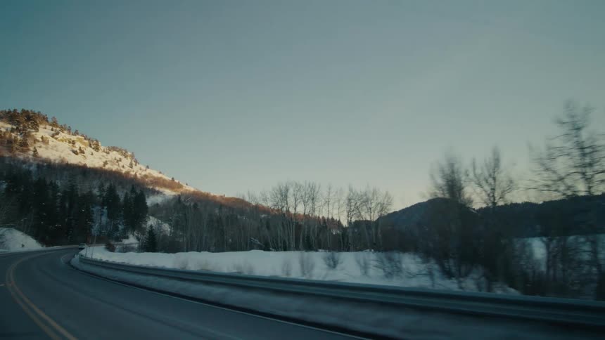 A roadway along a snow covered lake with mountains in the background and a white car traveling on the road