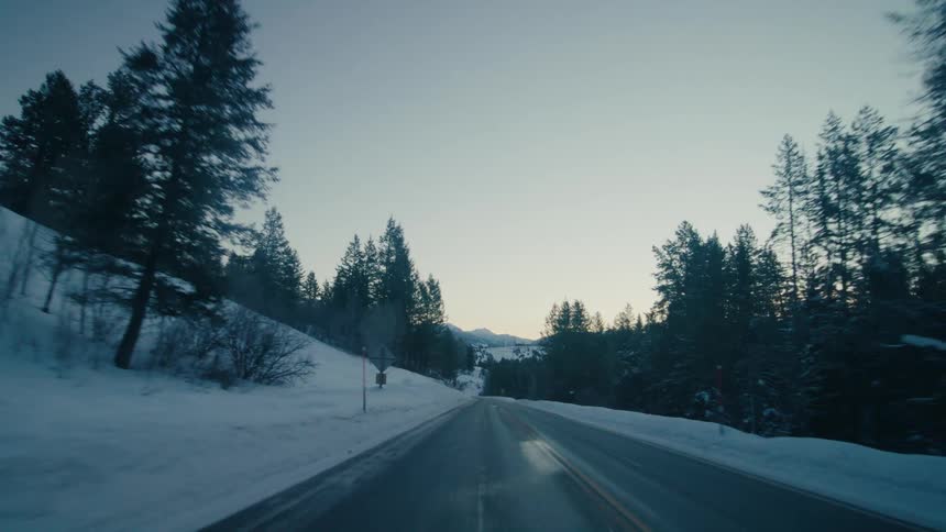 A roadway with snow covered roadsides with pine forests on either side and mountains in the distance