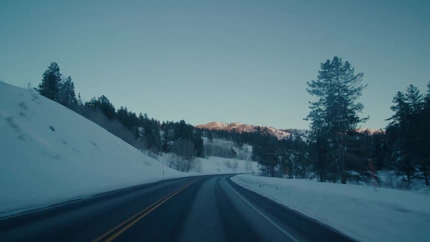 Road surface contrasts with snow as road passes conifer covered slopes