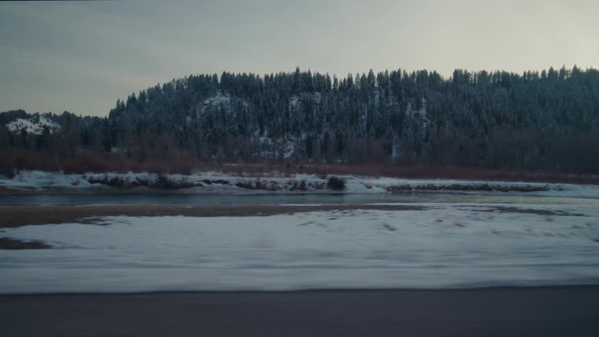 Traveling on a rural highway with snow and a stream along the roadside and forested mountains in the distance