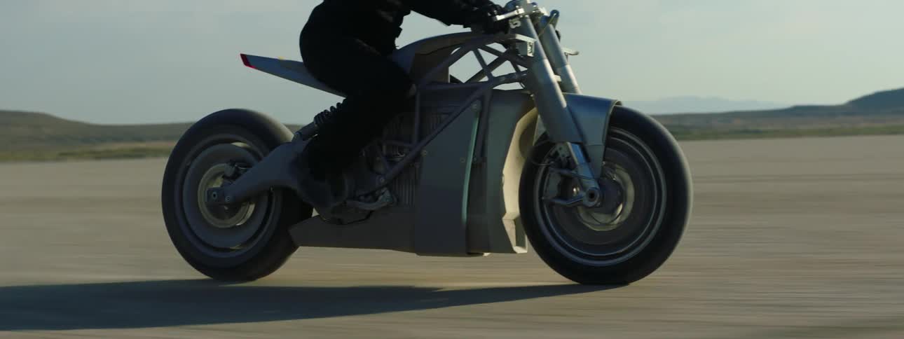 A person in black riding a motorcycle through the desert with mountains in the distance