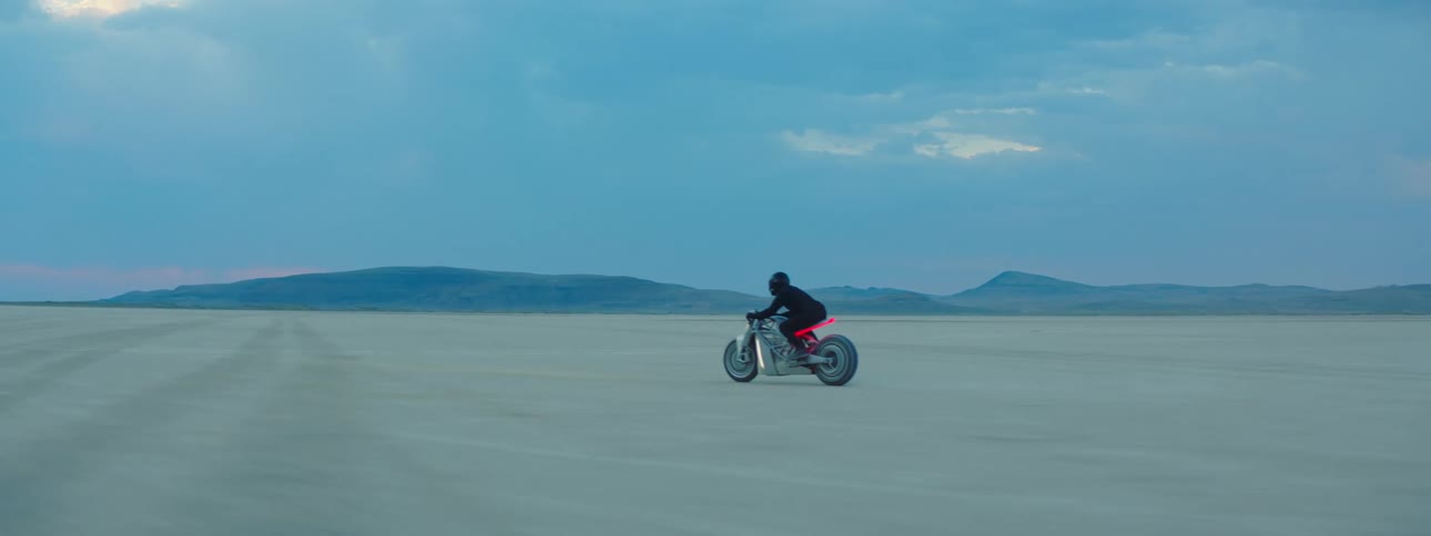 A person wearing all black driving a motorcycle through the desert with mountains in the far distance