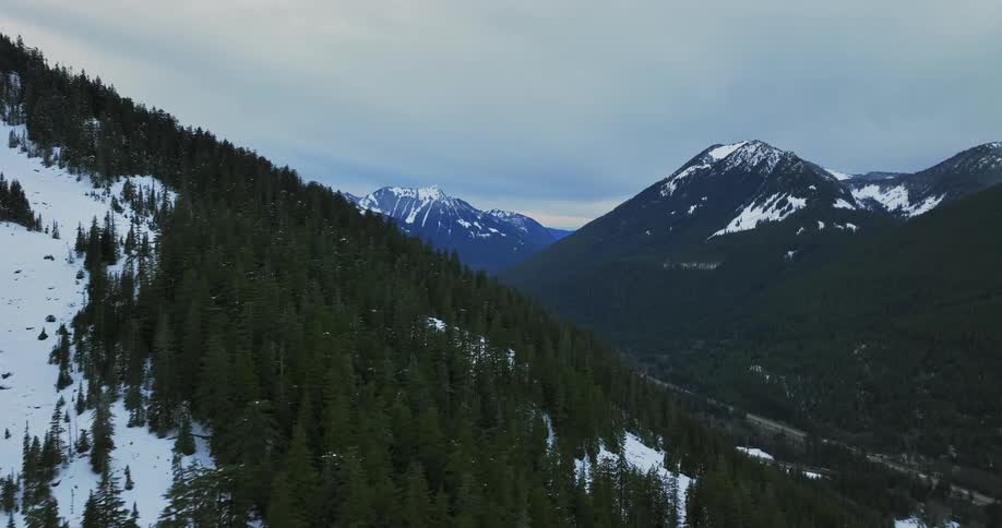 Traffic flows along road through narrow valley between snow and conifer covered mountains
