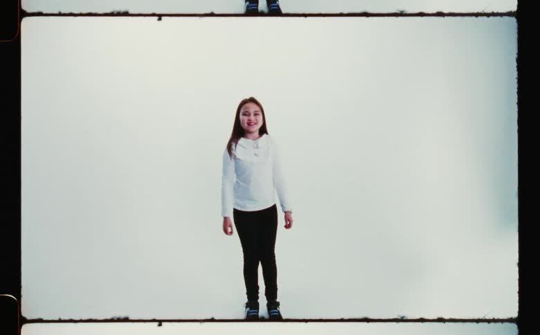 A little girl wearing a white sweater and black pants smiling and jumping up against a white backdrop