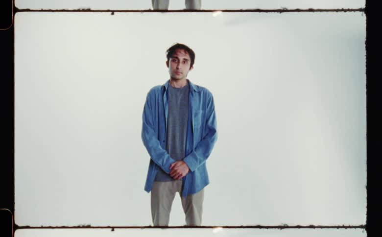 A man wearing a blue overshirt standing with his hands together in front of him against a white backdrop