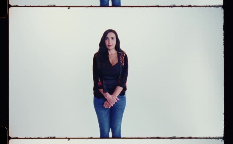 Woman with black wavy hair dark blue blouse and jeans poses for the camera in front of white screen backdrop in photography studio