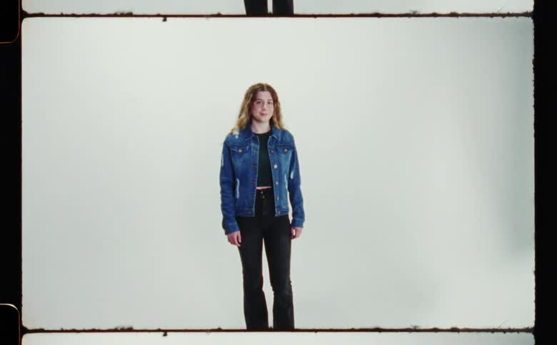 Woman with brown wavy hair denim jacket and black pantssmiles at camera in front of white screen backdrop in photography studio