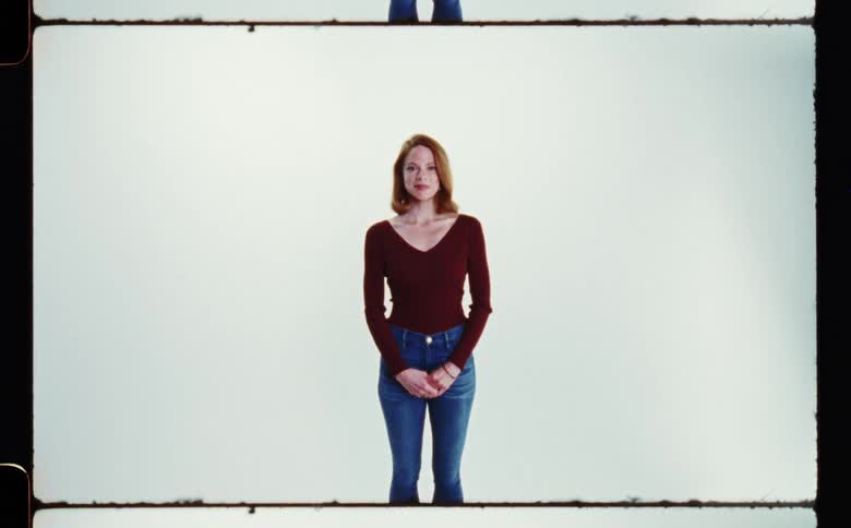 A red haired woman wearing a wide neck sweater and blue jeans is standing and smiling as she looks ahead