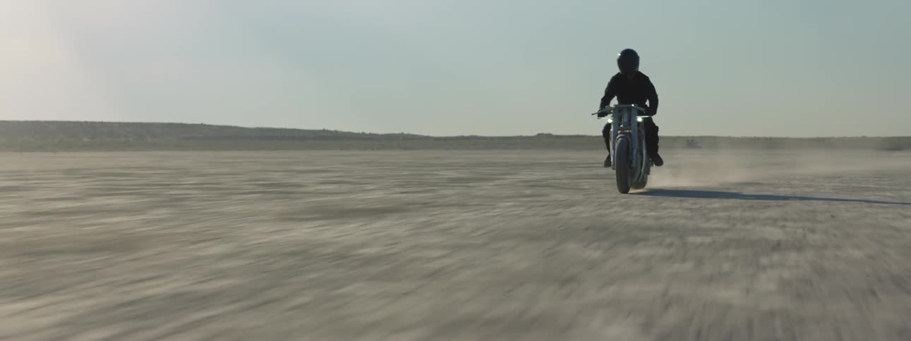 A motorcycle speeds across the desert leaving a trail of dust as the rider is wearing a black jacket and helmet