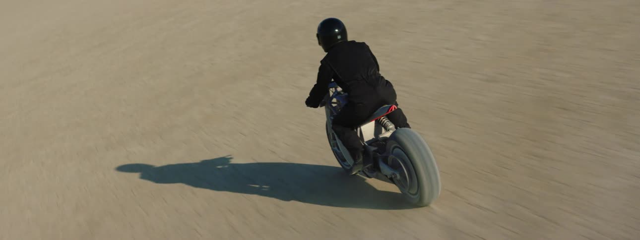 A rider wearing a helmet and dressed in black races a motorcycle over cracked packed sand in a desert
