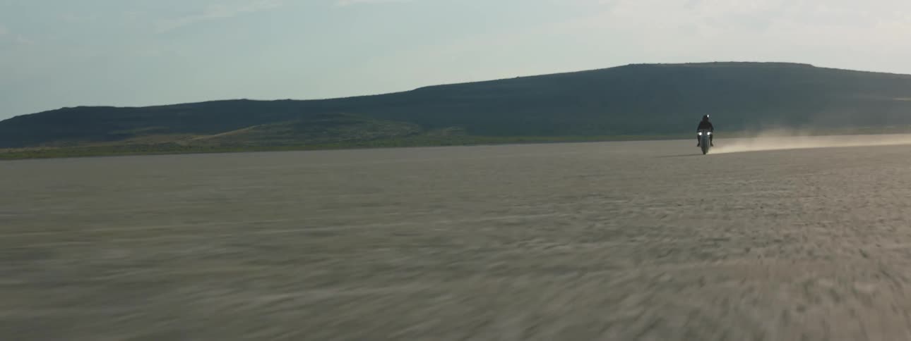 A motorcycle rider is racing across flat desert land with mountains in the background