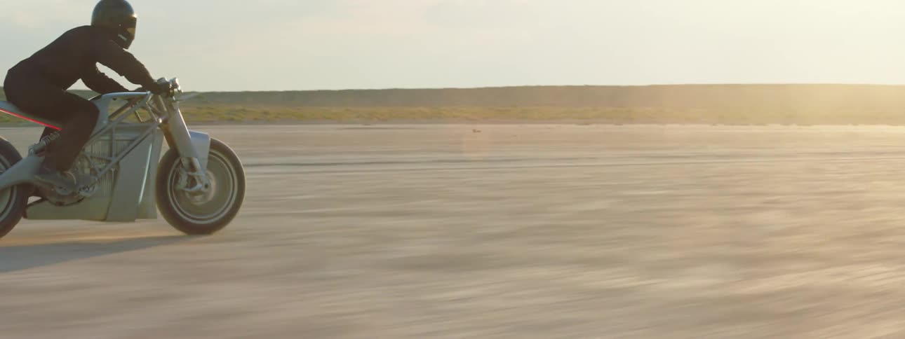 Biker with helmet with black jacket and pants rides silver motorcycle in the desert at dusk