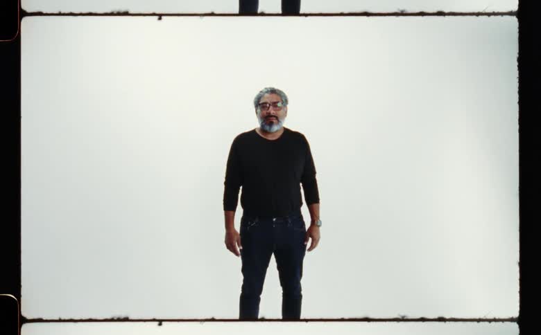 Man with gray beard glasses and black clothing poses for the camera in front of white screen backdrop in photography studio