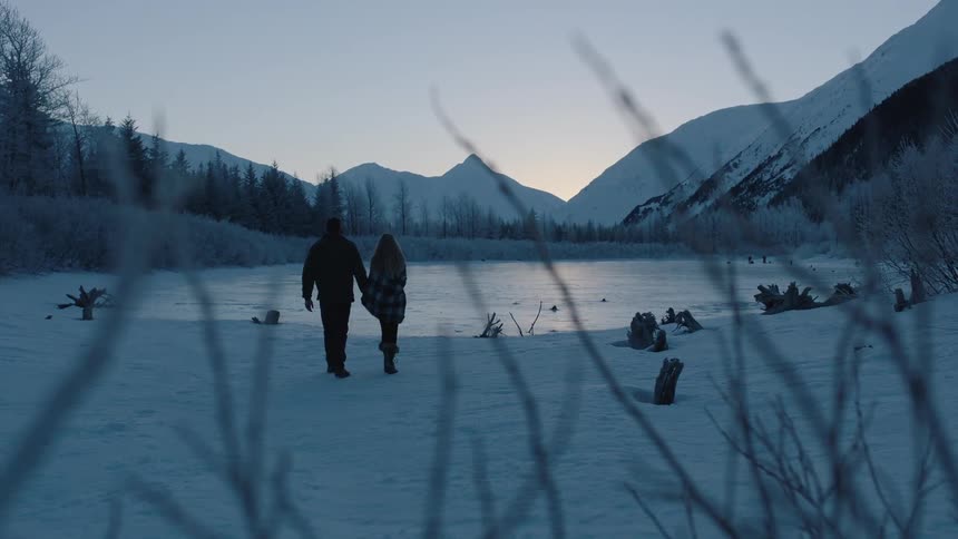 A man and woman snuggle and hold hands as they walk in a snow-covered landscape