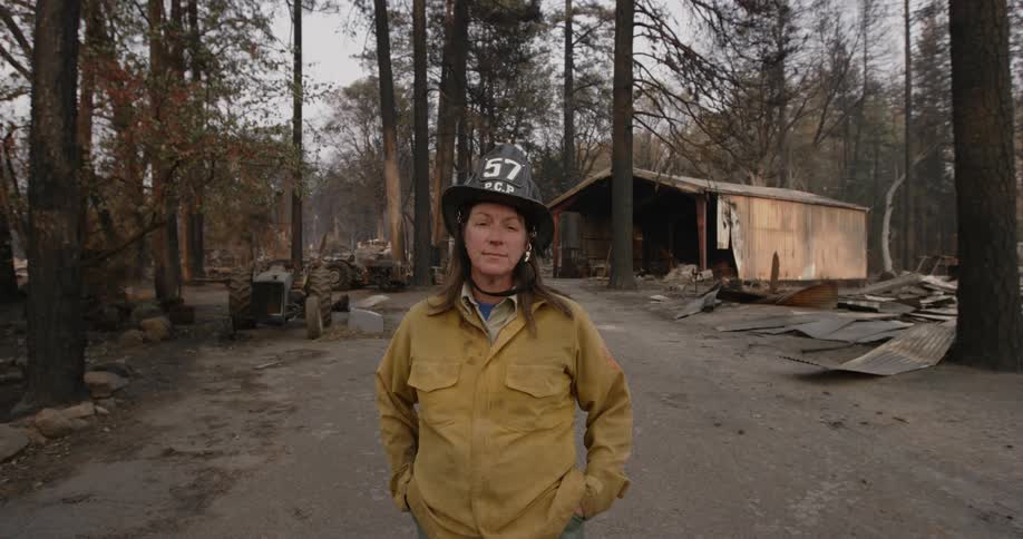 A woman fire fighter is standing outside building and tractors in the woods burned by the Paradise fire with sheets of torn metal siding on the ground