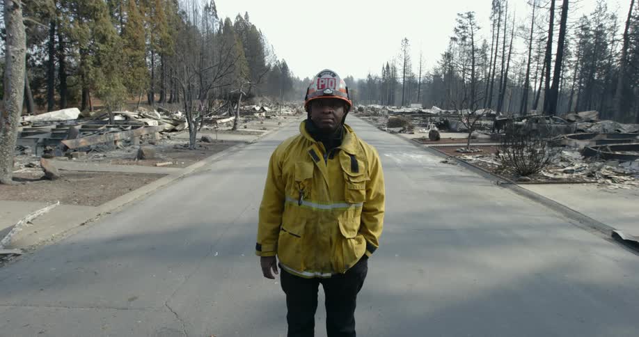 A fireman is standing on a street with driveways but the houses have been burned to the ground with rows of trees standing behind their remains