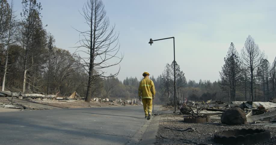 Fireman wearing a yellow suit and helmet walks around a burnt out neighborhood