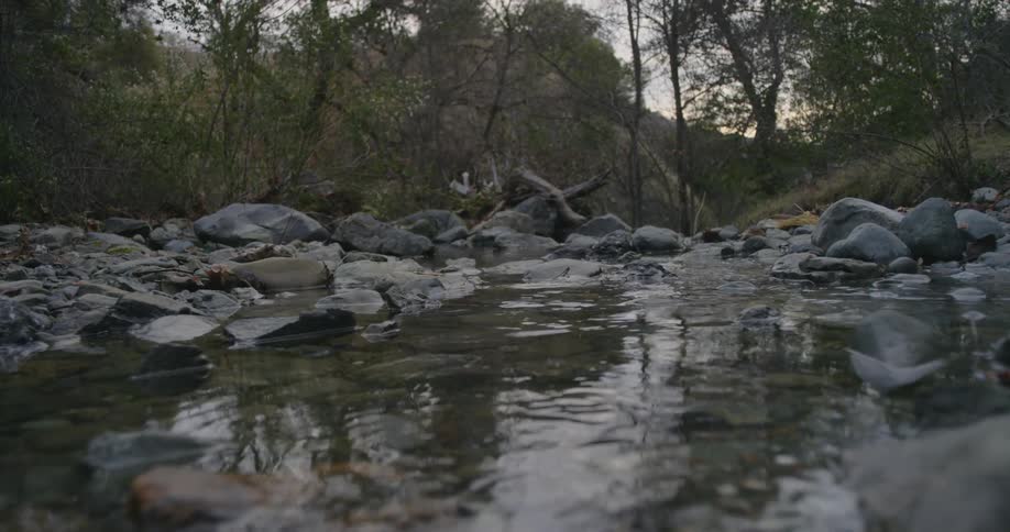 Two people wearing camouflage pants and boots step on rocks to cross over a stream in the woods