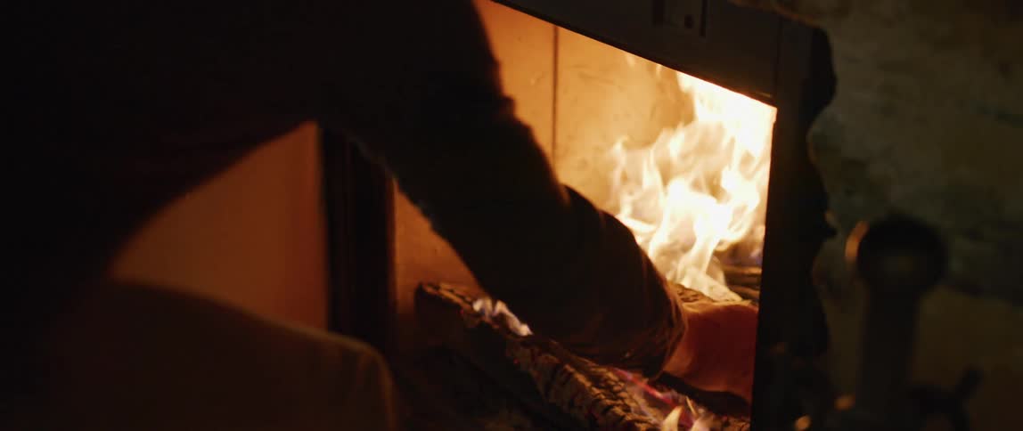 A man opens the door of a wood burning furnace and places another piece of wood on the fire