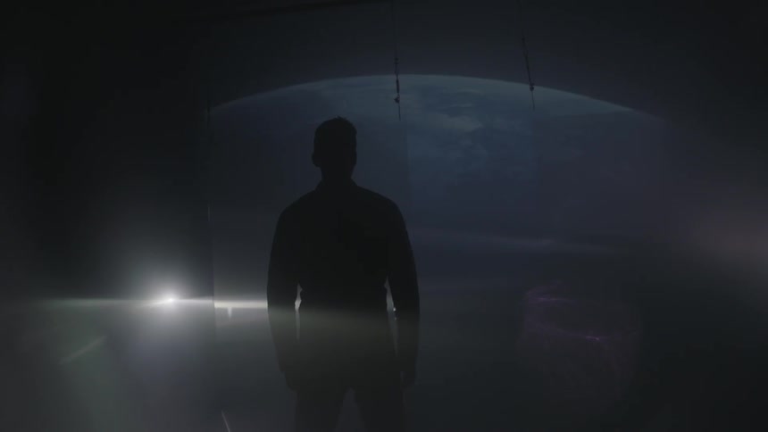 A man stands in a dark room as lights are projected around him