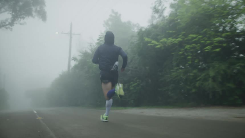 A jogger wearing shorts and a dark hoodie is running on a foggy rural road near trees