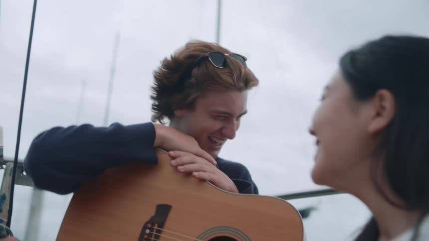 Man and woman sitting on sailboat with guitar laugh as they talk