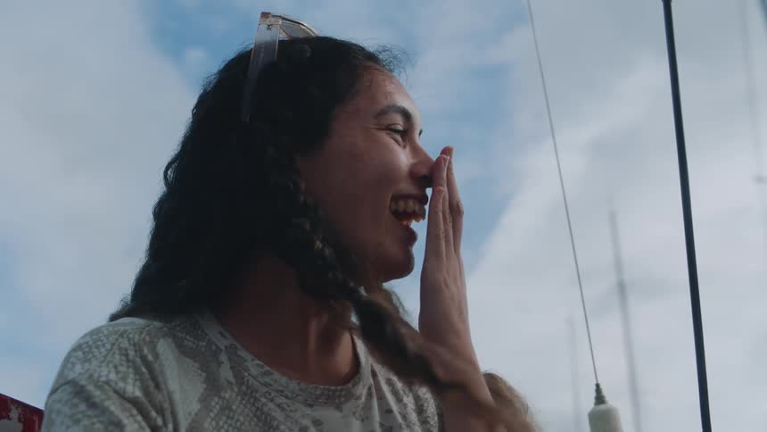 A girl smiles wide with her teeth showing as she places her hand in front of her mouth fingers straight up while she is laughing and rocking gently in a boat