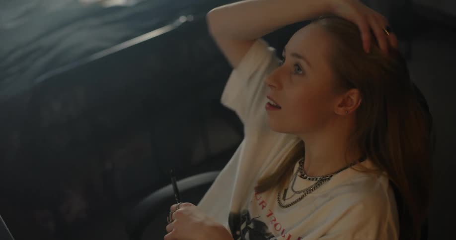 Blonde woman with necklace and white t shirt sits in armchair touching her hair and looking up in bedroom