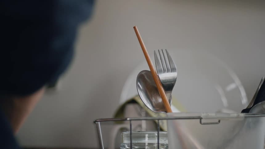 A metal dish drying rack with a spoon and a knife and a couple of other items is having dishes added to it by a person standing in front of the sink beside it washing them