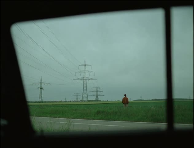Window of car parked near road shows man standing in field near transmission towers on overcast day
