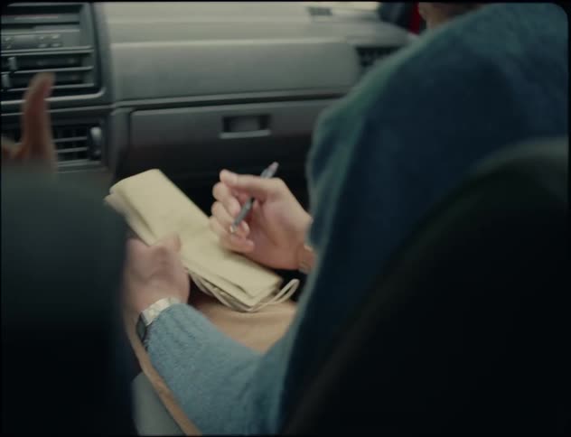 A driver and a passenger are traveling at a steady speed while the man is writing and the driver as snapping to get his attention which causes him to look playfully defensive and the aggressiveness of what the driver is saying